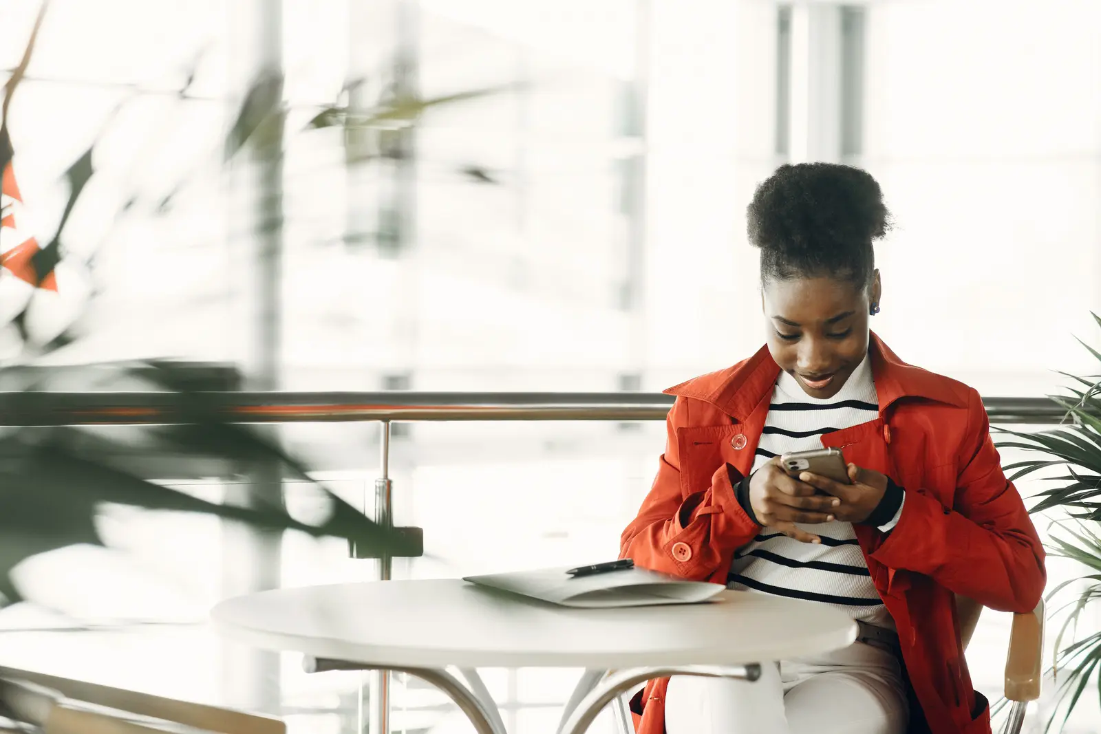 A woman at a cafe table using a phone, plants in the foreground.