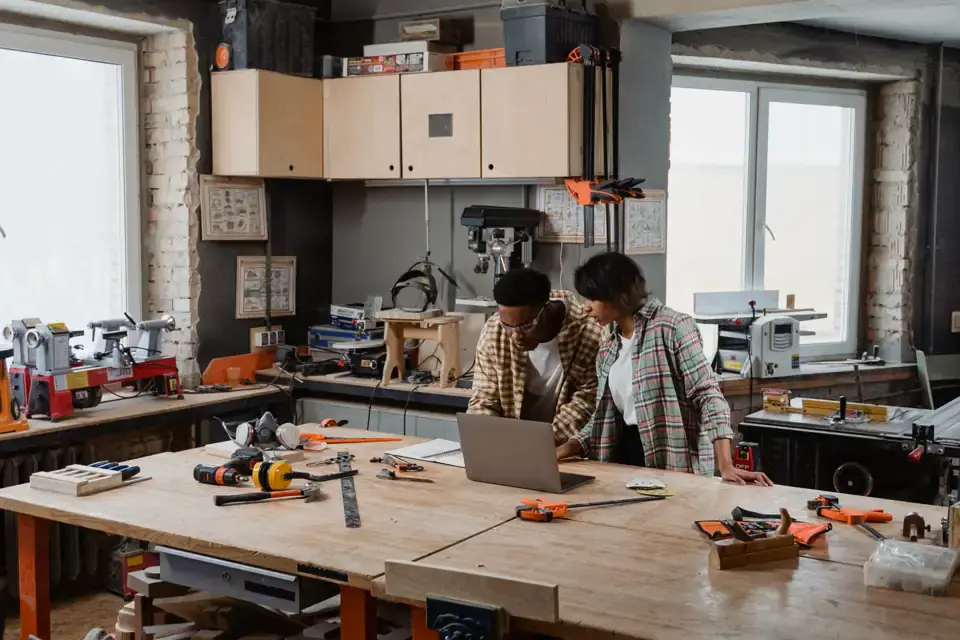 Two people at a workbench with a laptop in a workshop.